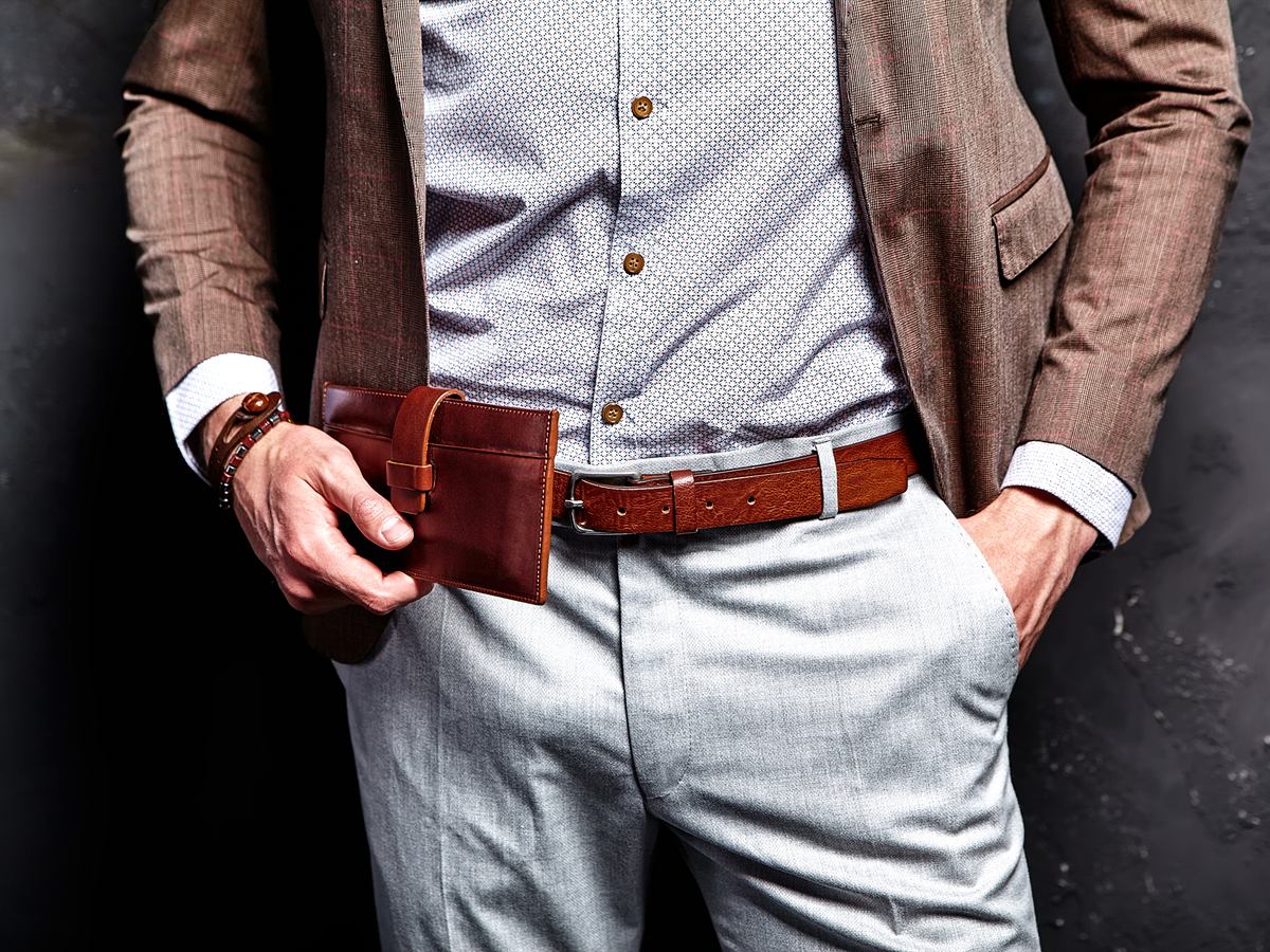 A man in a brown check jacket holding a brown leather wallet and wearing a tan leather belt